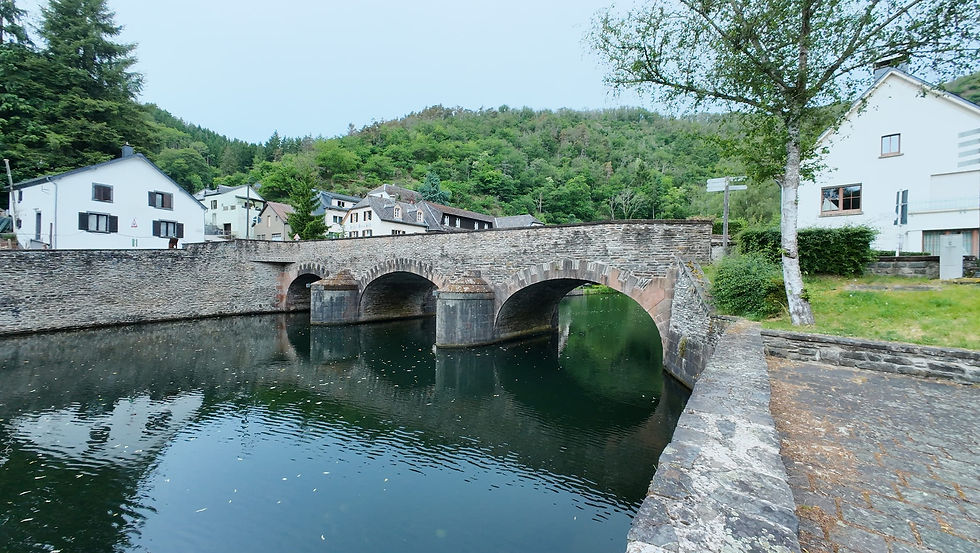 Steinbrücke mit drei Bögen über ruhigem Fluss, weiße Häuser und grüne Hügel im Hintergrund.