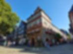 Historisches Fachwerkhaus an sonnigem Platz, Passanten spazieren, blauer Himmel, bunte Fensterläden und Blumen.