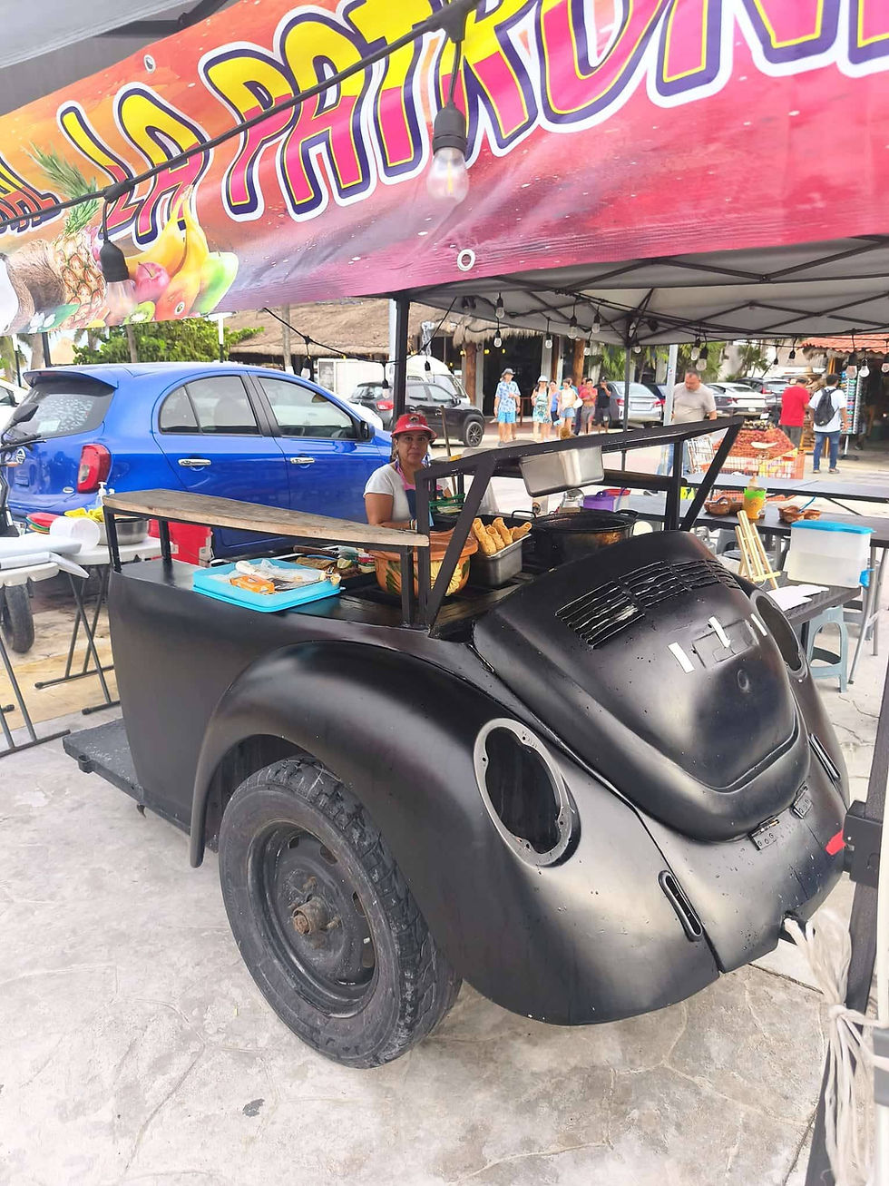 Verkäuferin an schwarzem Käfer Imbisswagen in Puerto Morelos unter Zelt, darüber buntes Schild; im Hintergrund geparkte Autos und Passanten.