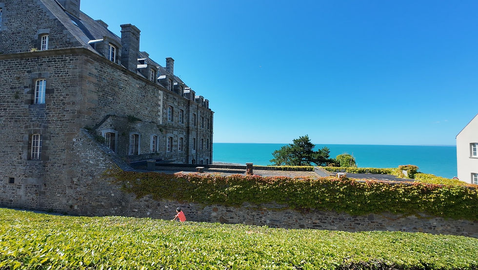 Historisches Steingebäude am Meer, umgeben von grüner Hecke. Sonniger blauer Himmel. Person im orangefarbenen Shirt geht spazieren. Granville Altstadt