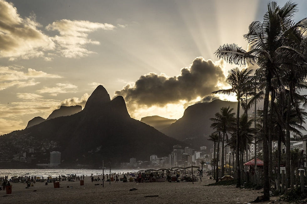 Strand bei Sonnenuntergang mit Menschen, umrahmt von Palmen. Die Silhouette eines Berges hebt sich vor einem strahlenden Himmel ab. Stimmung: ruhig. Rio de Janeiro