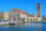 Steinkirche mit rotem Dach am Wasser, daneben Glockenturm. Blauer Himmel, griechische Flagge, Menschen spazieren entlang des Kais.