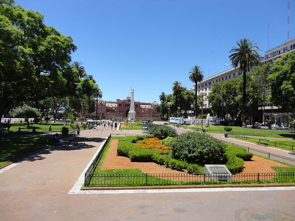 Weitläufiger Platz mit Palmen und Blumenbeeten, Menschen spazieren. Im Hintergrund ein rosafarbenes Gebäude und ein Denkmal bei blauem Himmel. Reiseziel Argentinien