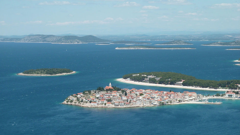 Insel mit roten Dächern und grünen Bäumen umgeben von blauem Meer; im Hintergrund weitere Inseln unter blauem Himmel. Ruhige Atmosphäre. Reiseziel Primošten, Kroatien