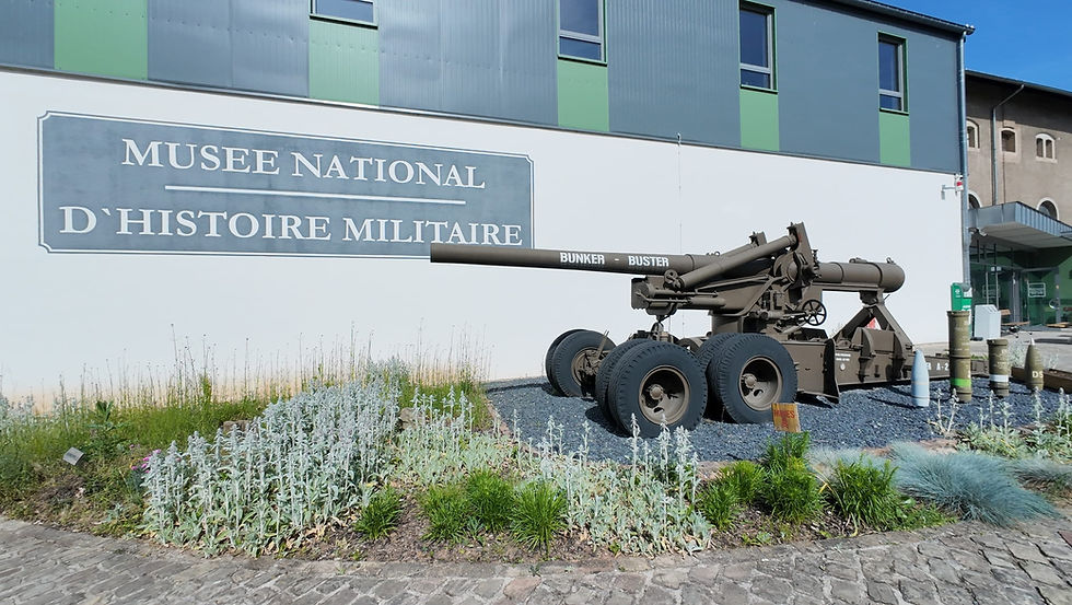 Military cannon display outside Musee National D'Histoire Militaire, with BUNKER-BUSTER shell on a museum wall under blue sky.