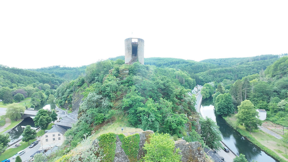 Burg-/Turmruine auf bewaldetem Hügel über Fluss und Tal, umgeben von grünen Bergen und kleinen Gebäuden im hellen Licht.