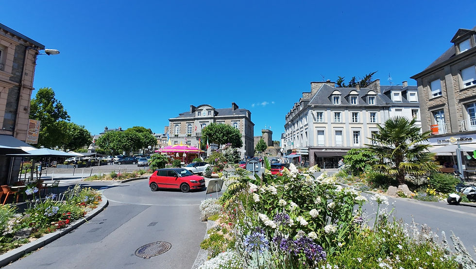 Kreisverkehr mit Blumenbeeten, rotes Auto, Gebäude im Hintergrund, blauer Himmel. Café mit rosa Schirmen, lebendige Stimmung. Reiseziel Avranches, Frankreich