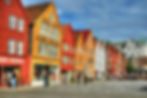 Bunte Holzhäuser am Hafen mit Passanten, Geschäften und Schildern; sonniger Platz unter blauem Himmel.