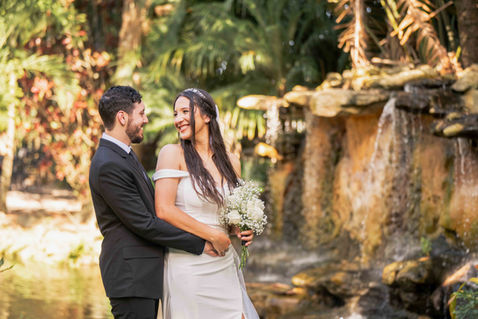 Portrait of a bride in a white dress holding a bouquet and a groom in a black suit with his arms around her. They're smiling at each other, with the forest and water in the background.
