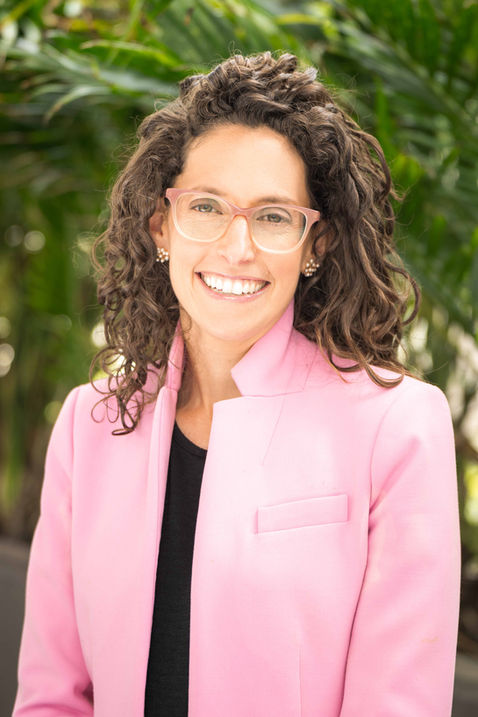 A portrait of woman wearing pink glasses and a pink blazer smiling in front of an outdoor backdrop.