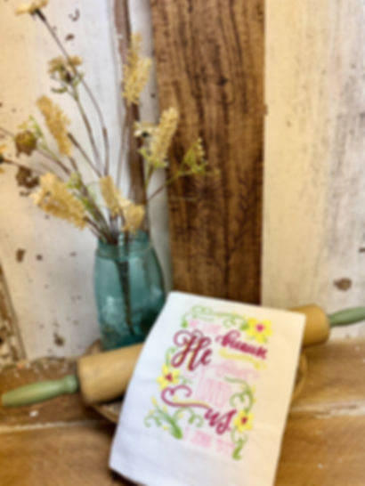 White cotton tea towel embroider with the verse "we love because He first loved us" John 4:19