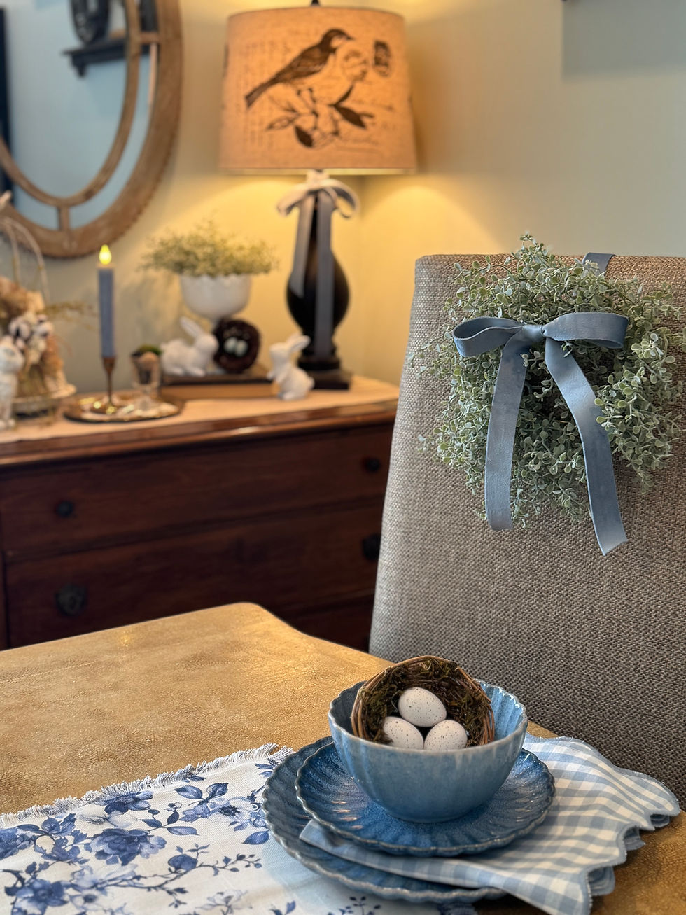 A cozy dining setup with blue dishes, faux eggs in a nest, and floral fabrics. A chair adorned with greenery and a lamp with bird art.