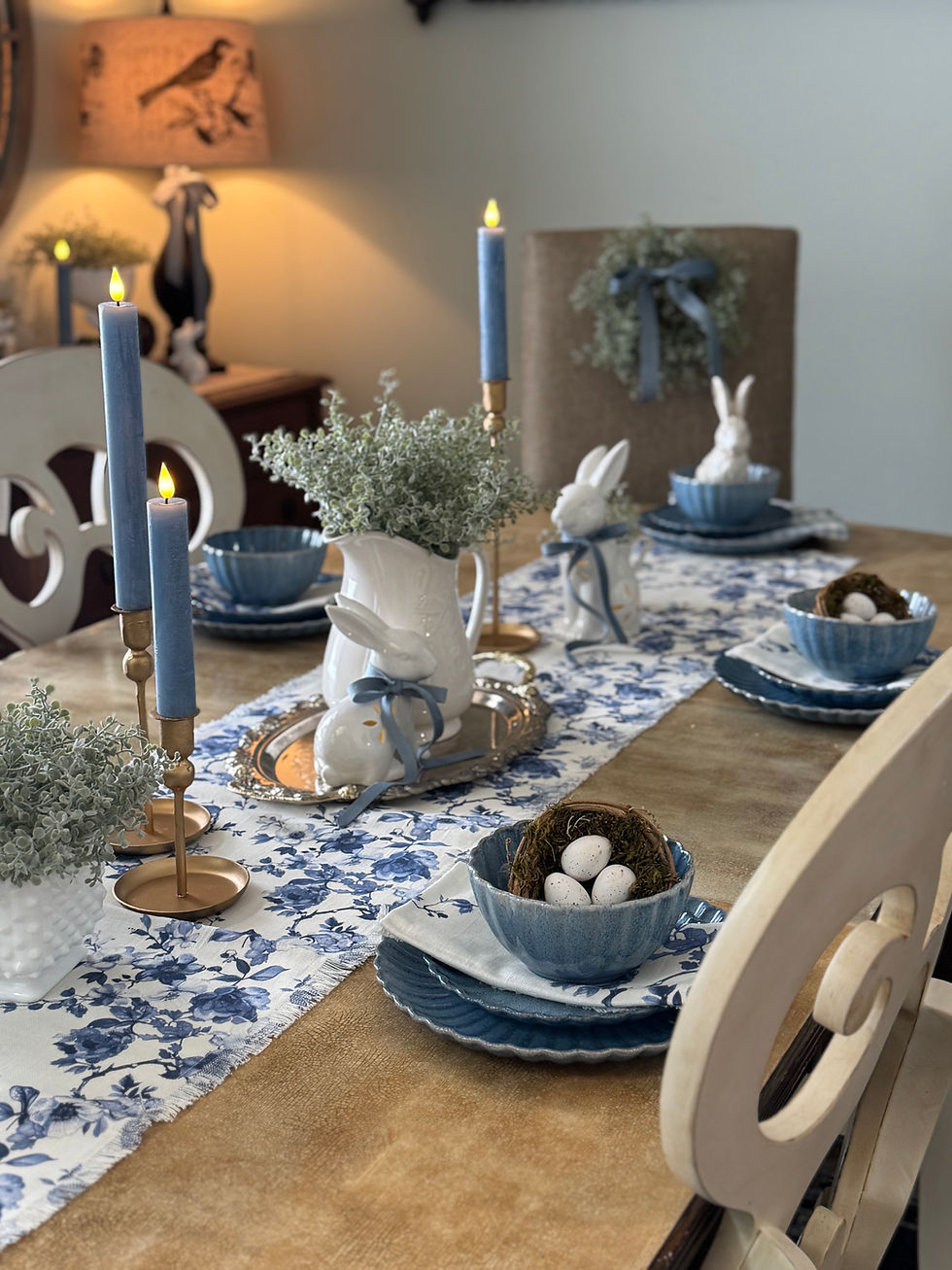 Elegant table setting with blue candles, rabbit figurines, floral runner, and blue dishes. Eggs in a nest centerpiece. Warm, cozy ambiance.