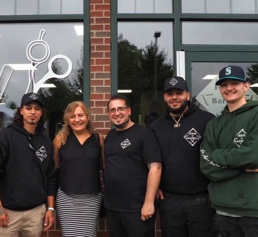 A group photo of the House of Barbers team standing outside their shop, smiling, with the shop's logo in the background.