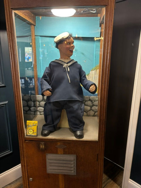 A sailor puppet in a blue uniform and white hat in a wooden display case. The background is painted with a seaside mural.