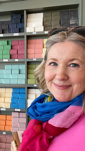 Blonde woman with colorful scarf standing in front of a wall of colorful soaps