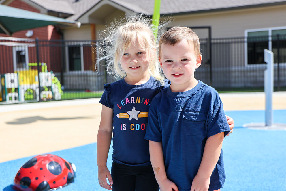 Girl and boy smiling, girl's shirt: "LEARNING IS COOL", outdoor playground.