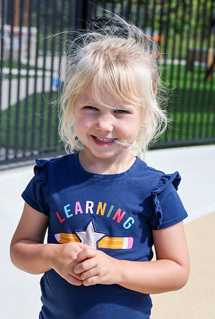 Blonde girl smiles, wearing "LEARNING" shirt on a sunny playground.