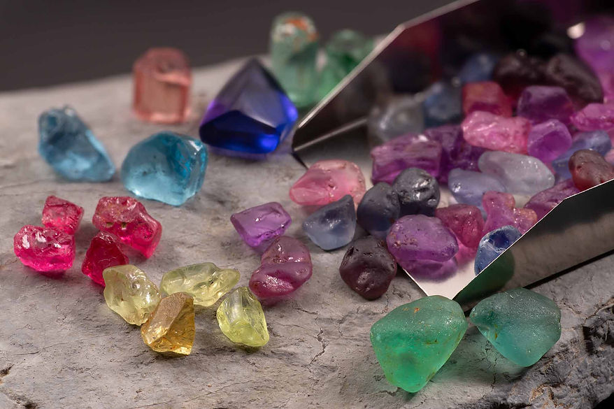 Assorted gem rough, spilling from a gem scoop. Includes sapphire, tanzanite, aquamarine, spinel, tourmaline, and chrysoberyl