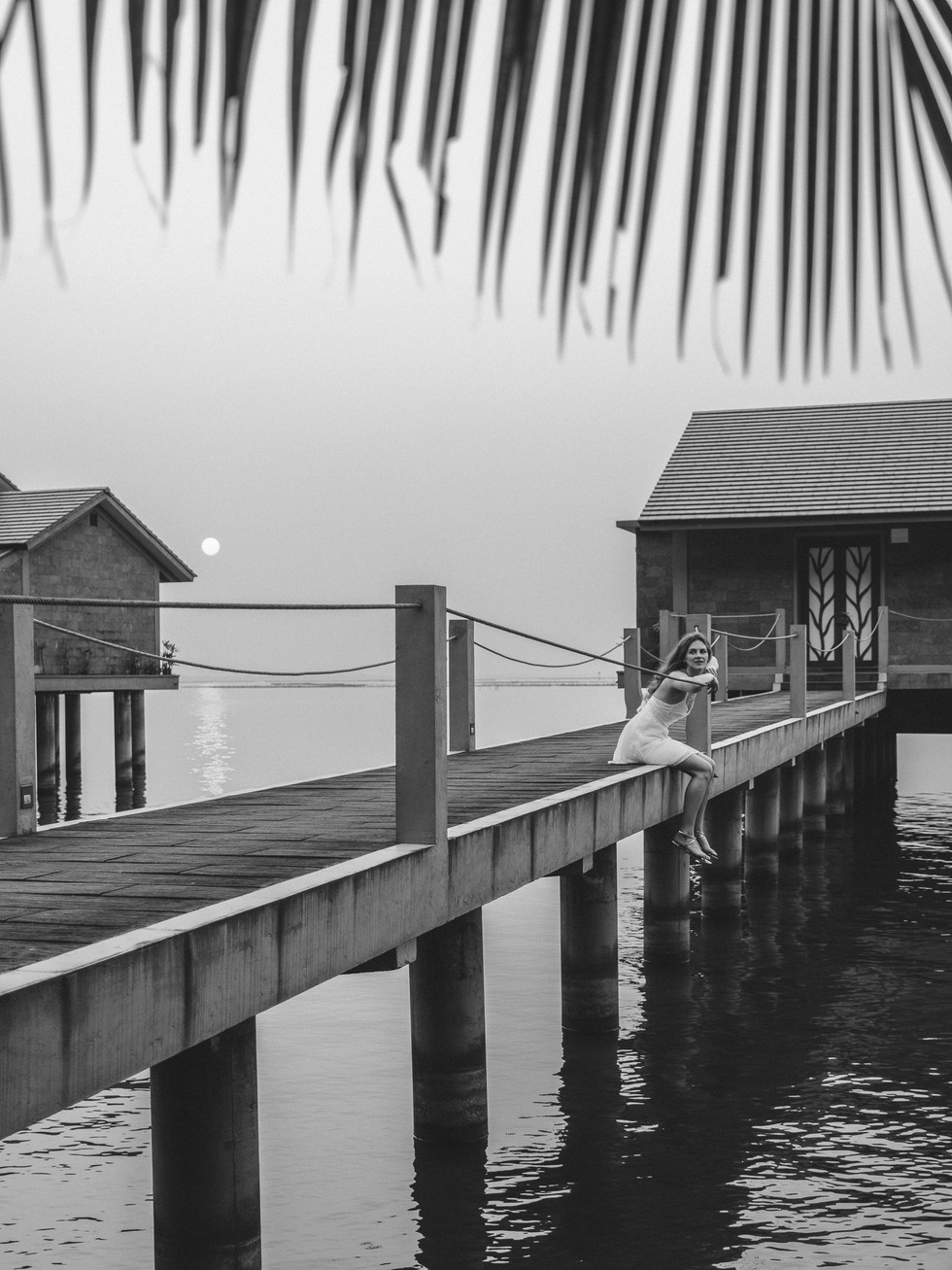junge Frau sitzt bei Sonnenuntergang auf einem Steg zur Wasservilla und schaut in die Kamera, Hue, Vietnam