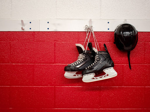 Hockey skates and a helmet are hanging from hooks on a white and red cinderblock wall.