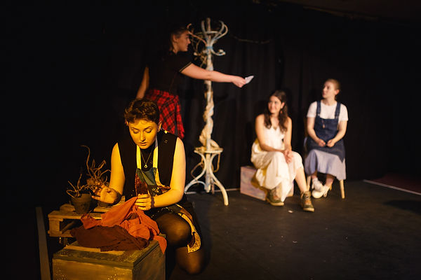 In the foreground there is a woman kneeling beside a wooden box, handling some red fabric. She is wearing a patchwork apron and a black dress. In the background, out of focus behind her there is a woman in a black top and red flannel handing a white envelope to the two women on the far right. There is a wooden coat stand between the woman standing and the two sat down. The woman on the far right is wearing a white top, denim apron and blue skirt with white sneakers. The woman to her left is sat on a pale box and she is wearing a white dress and green hiking boots.