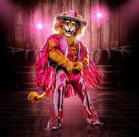 Löwenkostüm mit rosa Hut, Umhang und glitzerndem Anzug auf Bühne.