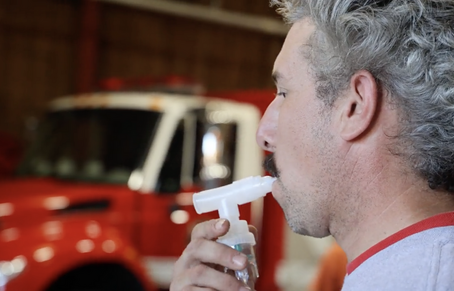 A firefighter with curly gray hair stands in a fire station bay, using a white nebulizer mouthpiece for a glutathione treatment. A red fire engine is visible in the background, slightly out of focus.