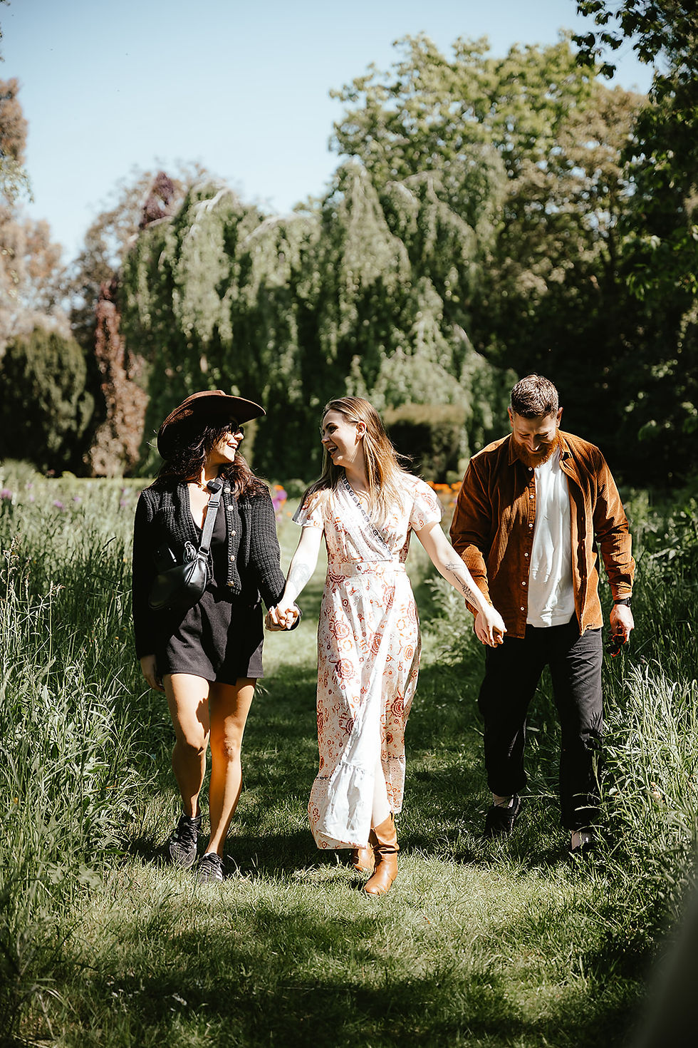 Three people walk hand in hand on a grassy path, smiling. They are surrounded by lush trees, under a bright blue sky.