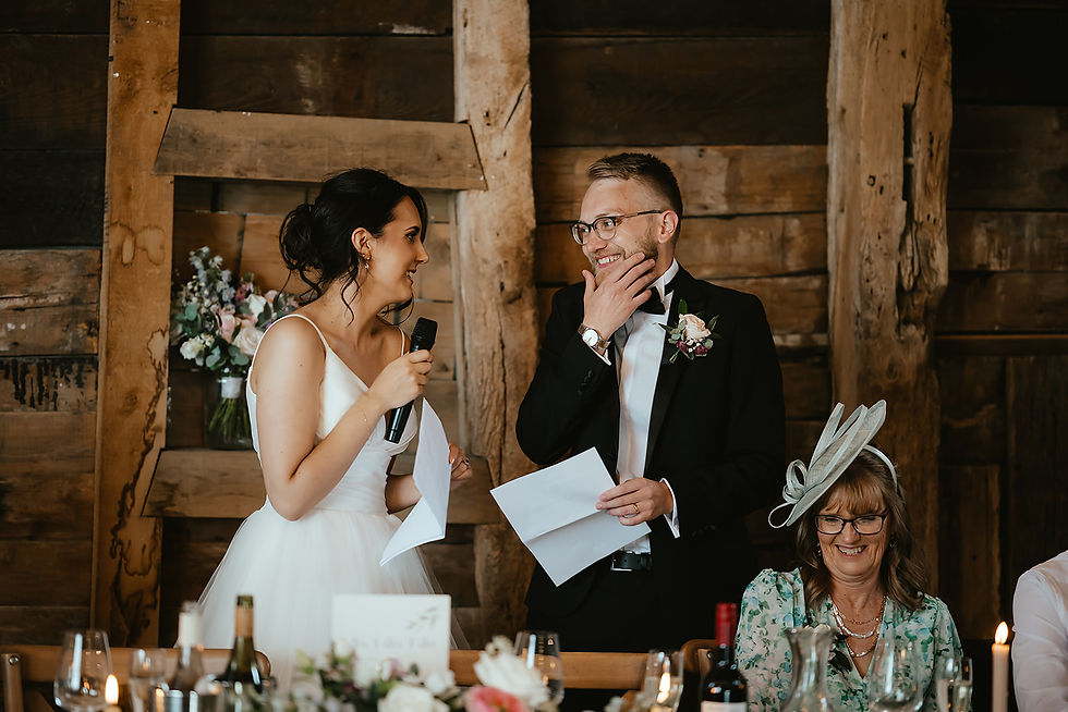 Bride and groom laughing during speech in rustic setting. Bride in white dress with a microphone; groom in suit holding paper. Candles and flowers decorate table.