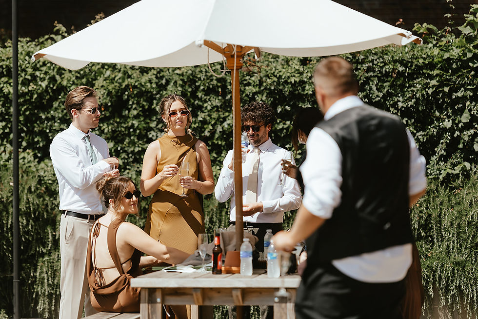 Guests in summer attire gather under a white umbrella at an outdoor table, chatting and holding drinks in a sunny garden.