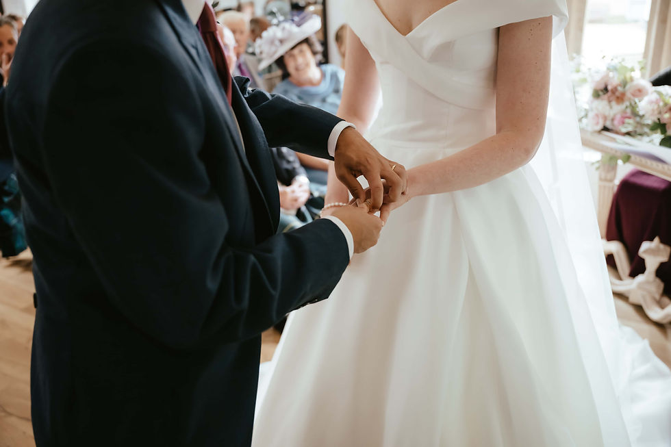 Bride and groom exchange rings during a wedding ceremony. The bride wears a white dress; guests observe in the softly lit background.