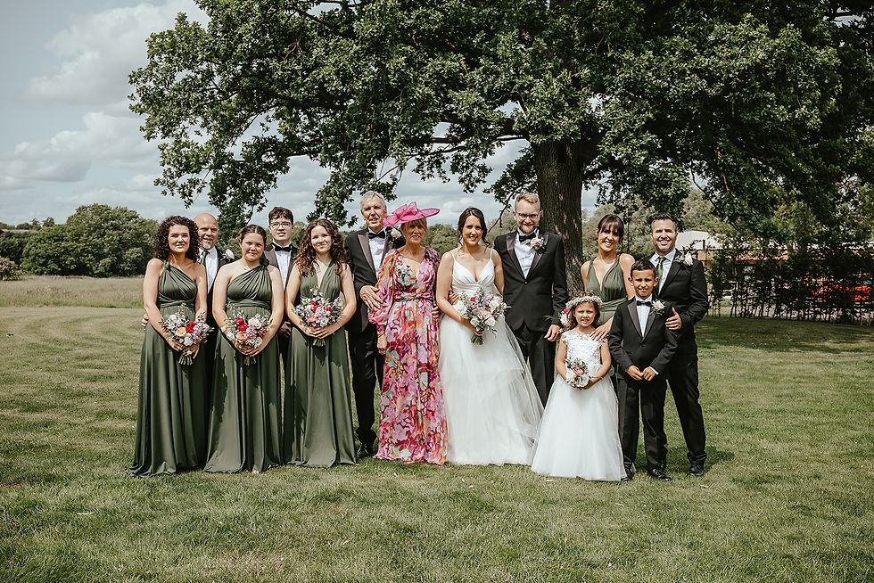 Wedding party poses in a garden, featuring people in dark green dresses and suits, a bride in white, and vibrant pink accents. Smiling faces.
