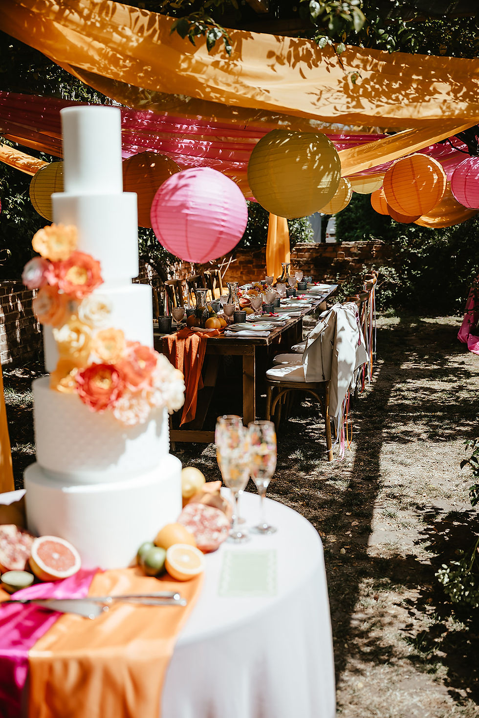Three-tiered cake with flowers on a table, surrounded by colorful lanterns and orange drapes. Sunny outdoor setting, festive mood.