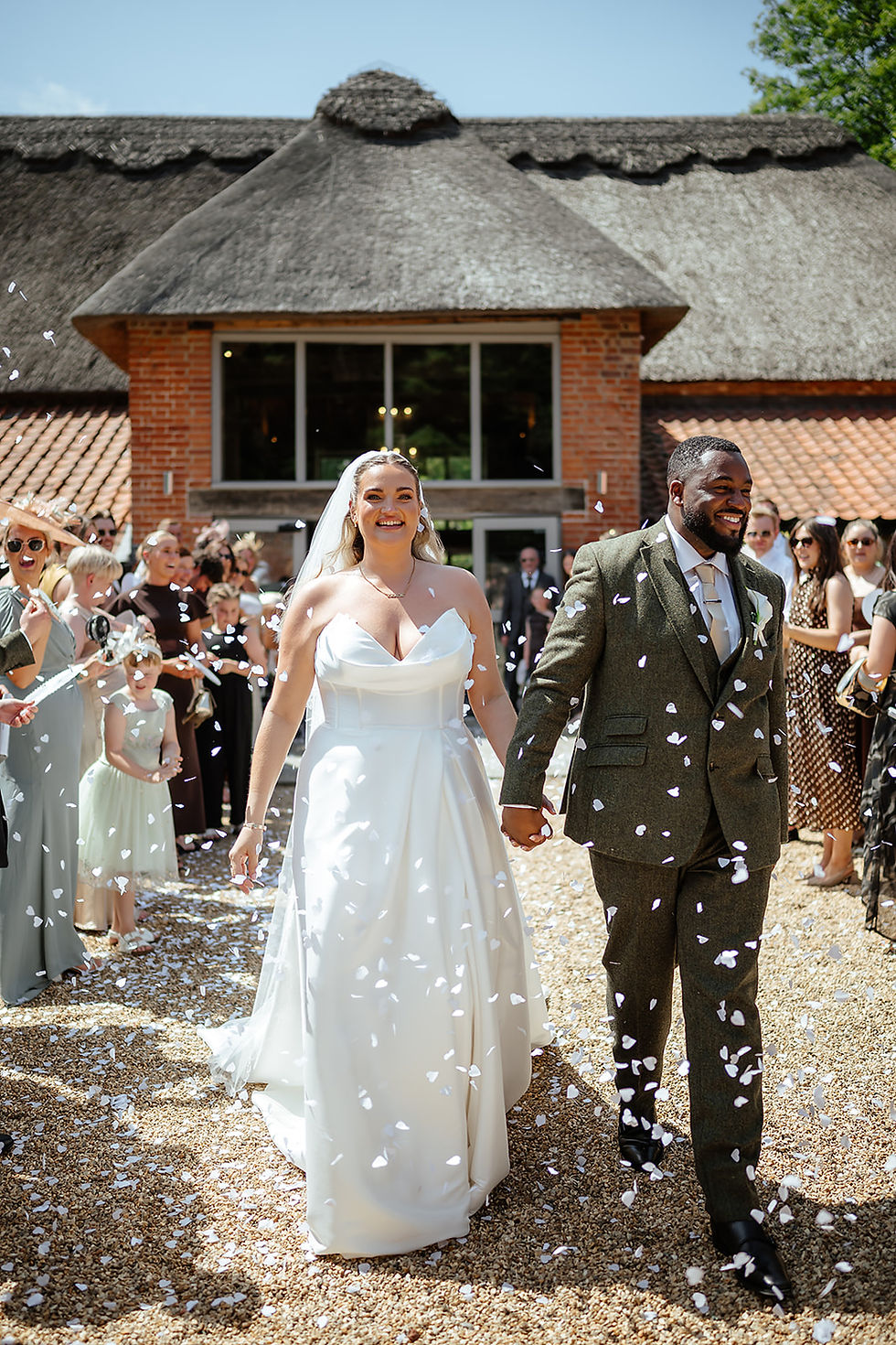 Smiling bride and groom walk hand in hand through confetti outside a thatched-roof venue, guests cheering.