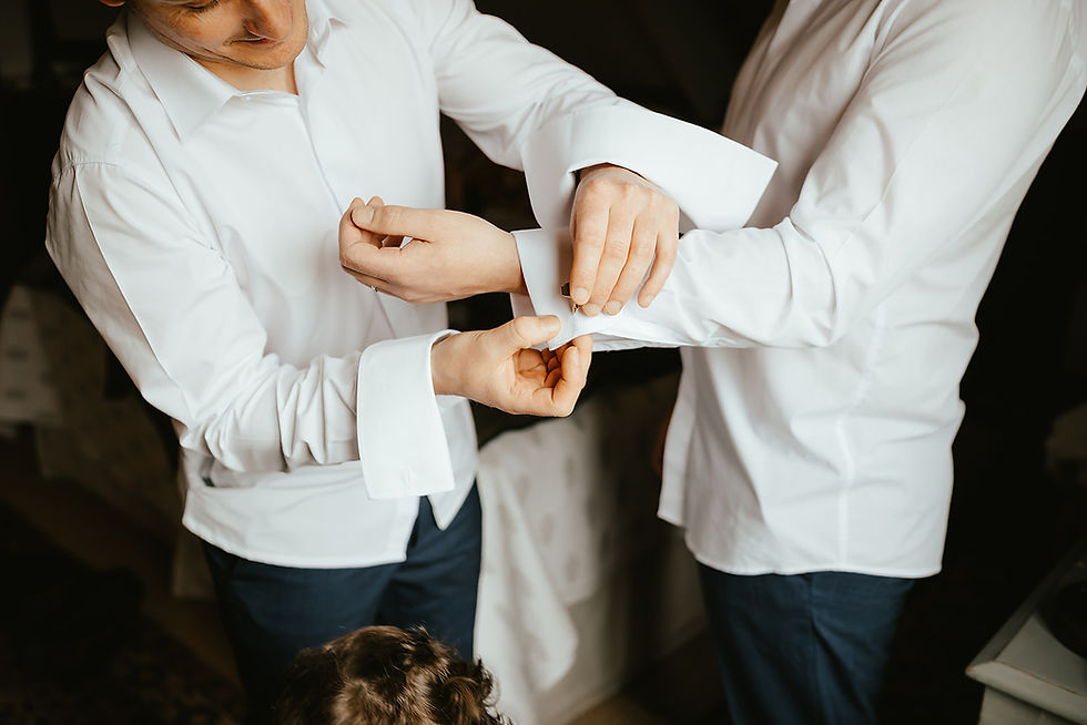 Two men in white dress shirts fasten a cufflink together in a dim room, with a child’s head visible at the bottom.