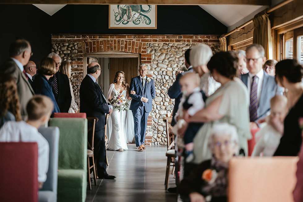 Bride in white dress and older man walk down aisle between seated wedding guests in a rustic stone hall, smiling.
