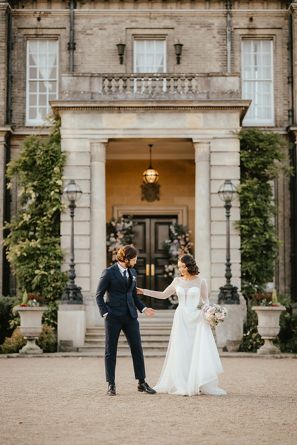 Bride in white gown and groom in navy suit joyfully walk outside an elegant building with floral decor, exuding a romantic mood.