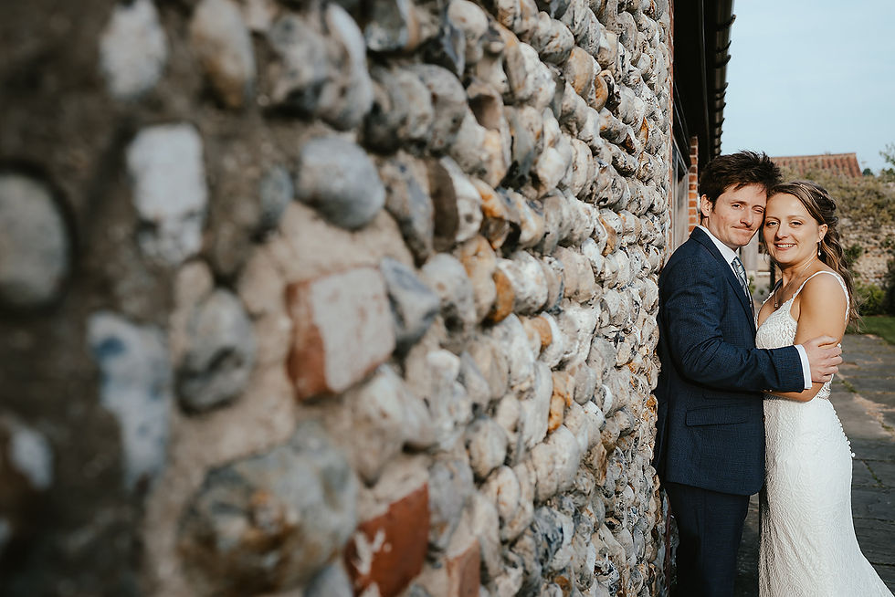 Bride and groom embrace beside a pebble stone wall outdoors, smiling softly in wedding attire.