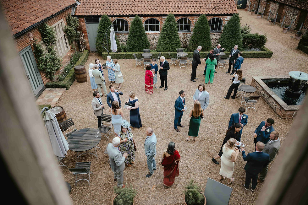 Guests in formal attire mingle in a courtyard with tables, greenery, and a fountain, set against a brick building with a tiled roof.