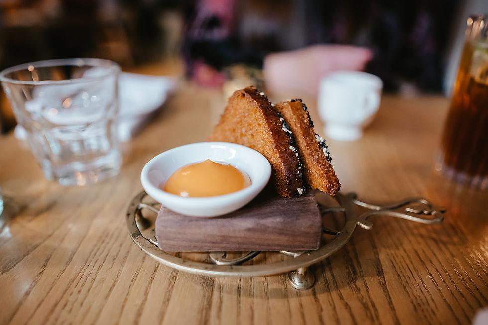 Fried triangles with sesame seeds sit on a decorative stand with dipping sauce on a wooden table. Nearby are a glass and a teacup.