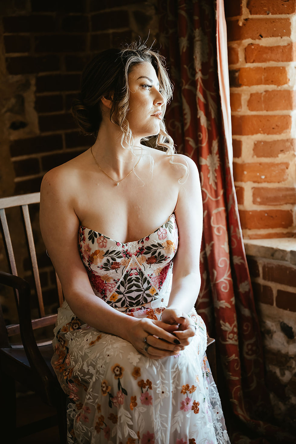 Woman in floral gown sits by a window in a rustic room with brick walls and red drapes, gazing thoughtfully outside.