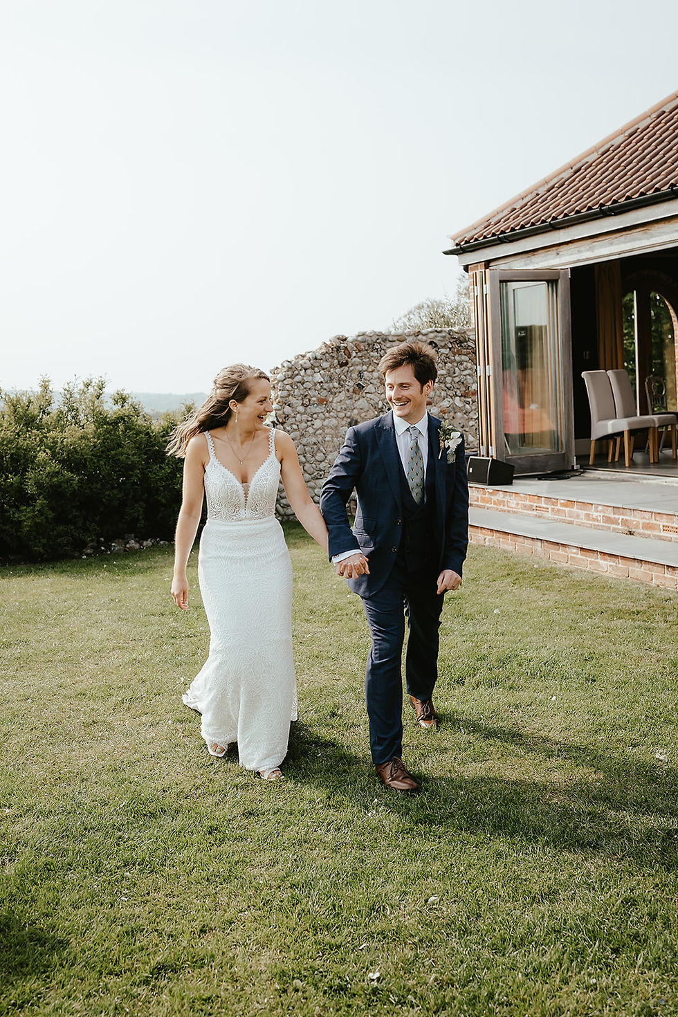 Bride in white lace dress and groom in navy suit hold hands and laugh while walking across a garden by a house