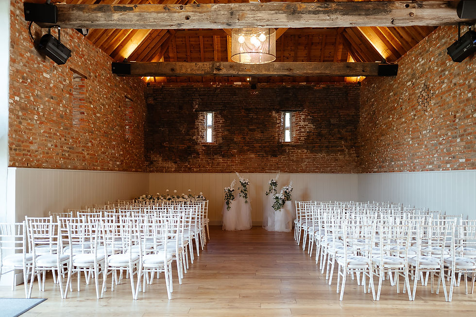 Rustic wedding venue with rows of white chairs, floral décor, exposed brick walls, and warm wooden beams under soft lighting