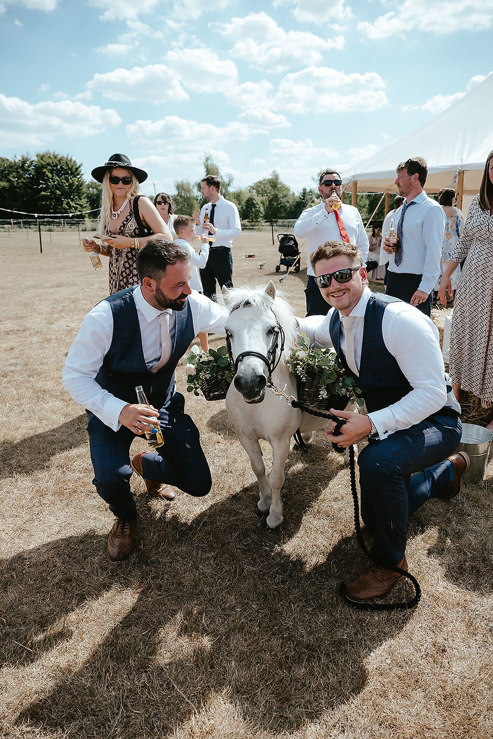 Two men in suits crouch beside a white pony with flowers. They're on a sunny lawn with others and a tent nearby, creating a festive mood.