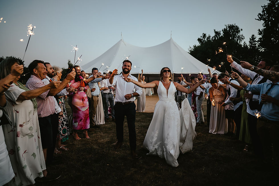Bride and groom walk through guests with sparklers, joyful and smiling, in front of a white tent at dusk. Celebratory and festive mood.