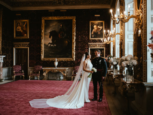 Bride and groom stand smiling in an ornate room with red carpet, gold-framed paintings, and chandeliers at Holkham Hall. Elegant and romantic setting.