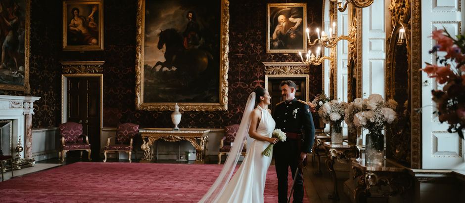 Bride and groom stand smiling in an ornate room with red carpet, gold-framed paintings, and chandeliers at Holkham Hall. Elegant and romantic setting.