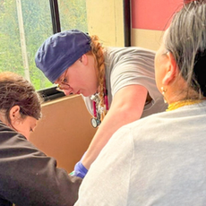 A nursing student wearing gloves and a stethoscope works alongside another healthcare team member while providing care to a seated patient in a clinic exam room. Large windows overlook a hillside community outside as the team focuses on the patient during a hands-on healthcare visit.