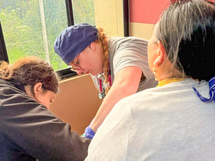 A nursing student wearing gloves and a stethoscope works alongside another healthcare team member while providing care to a seated patient in a clinic exam room. Large windows overlook a hillside community outside as the team focuses on the patient during a hands-on healthcare visit.
